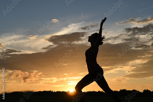Silhouette of slim young woman practicing yoga warrior pose, sunset in the background