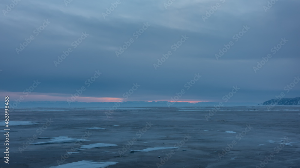 Twilight over a frozen lake. The cloudy sky is highlighted in pink. Patches of snow are visible on the ice. Baikal in winter