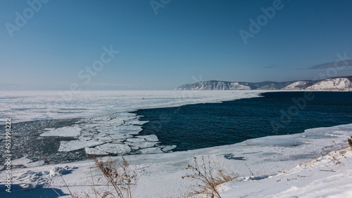 A non-freezing river flows out of a frozen lake. Blue water and white ice. Ice floes float on the surface. Snow and dry grass on the shore. Mountains against the azure sky.Baikal. Angara