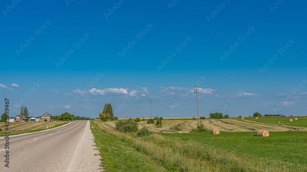 Road, mown field with hay rolls and electricity poles.