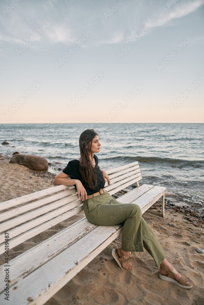 Beautiful brunette woman looking far into the sea horizon. Girls sits on white bench on the beach. Concept of work-life balance and wellbeing.