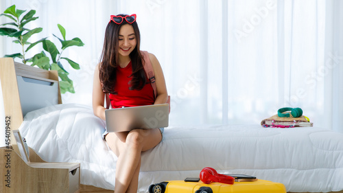 Young happy and excite Asian female traveler getting ready for a summer holiday trip vacation searching for travel information using laptop in bedroom