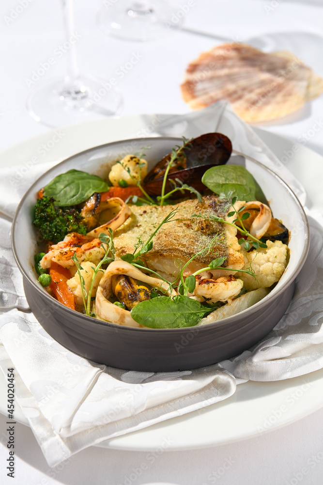 Elegant seafood stew with cod fish and vegetables in pot. Healthy fish dish - baked codfish, shrimp, mussels on broccoli and tomato. Serving white tablecloth and glass in summer day with shadow. Elegant seafood stew with cod fish and vegetables in pot. Healthy fish dish - baked codfish, shrimp, mussels on broccoli and tomato. Serving white tablecloth and glass in summer day with shadow.