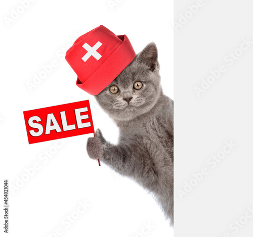Cat wearing doctors cap looks from behind empy white banner and shows sale symbol. isolated on white background