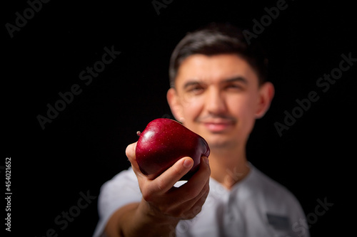 A young man who is a medical doctor in a white medical uniform and with red apple poses against a black background in the studio