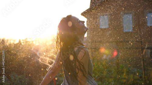 Joyful brunette girl enjoys her evening in the countryside by dancing in the rain. Stunning golden sun rays shine on playful young woman spinning and enjoying a spring shower. CLOSE UP, LENS FLARE