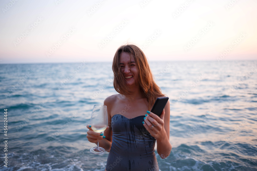 beautiful young woman with glass of wine and phone at sunset by the sea