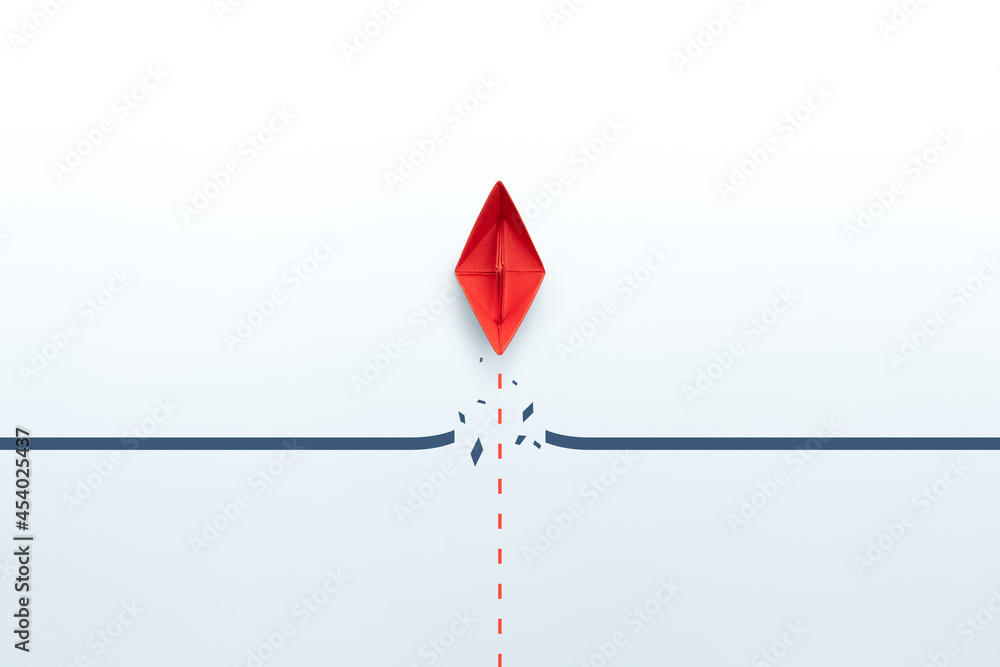 Concept of overcoming barriers, goal, target, with red paper ship breaking through obstacle on white background Concept of overcoming barriers, goal, target, with red paper ship breaking through obstacle on white background