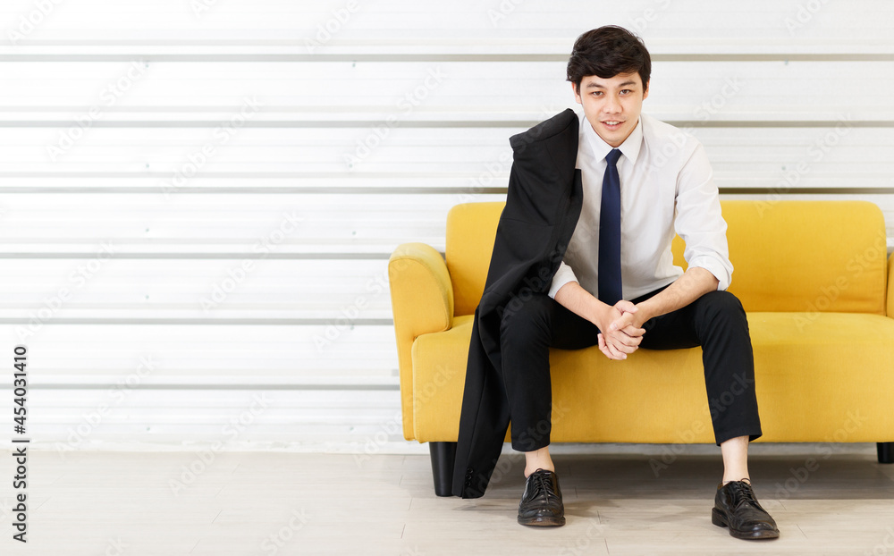 Fototapeta premium Horizontal portrait shot of young adult Asian handsome man in a white shirt with a suit on the shoulder sitting on the sofa and looking at the camera with hand to chin gesture posing in the studio