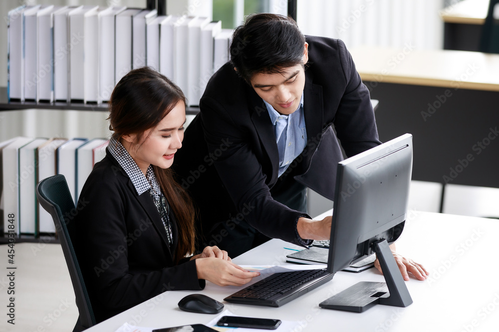 A smart young business woman wearing modern glasses working on computer together with a confident business young man standing next to her looking at the screen and discussing. Teamwork concept. A smart young business woman wearing modern glasses working on computer together with a confident business young man standing next to her looking at the screen and discussing. Teamwork concept.