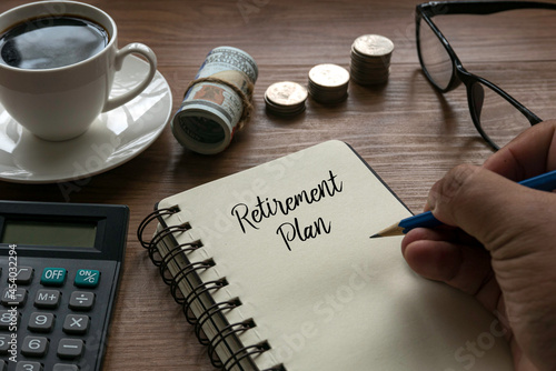 A hand writing a word ' Retirement Plan ' with a stack of coins, money, glasses, calculator and a cup of coffee on a wooden background.