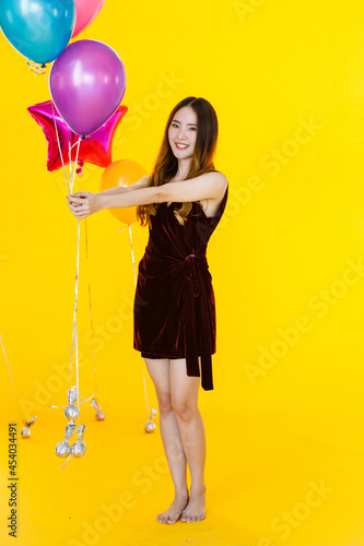 Portrait studio shot of Asian happy girl wears sexy dress standing posing smiling look at camera holding colorful helium air balloons in hand in celebration party in front of yellow wall background