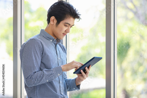 Portrait shot of Asian young happy handsome successful male businessman standing holding touch screen tablet in hands in front glass windows with blurred green tree background Free Download Portrait shot of Asian young happy handsome successful male businessman standing holding touch screen tablet in hands in front glass windows with blurred green tree background - GFXLoad