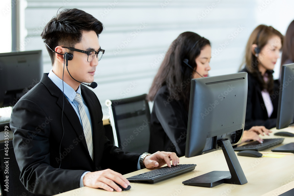 Cheerful Asian man in headset smile with cheerful and happy while working on computer at desk with female coworkers in help desk office. Sevice mind and ready for marketing support concept Cheerful Asian man in headset smile with cheerful and happy while working on computer at desk with female coworkers in help desk office. Sevice mind and ready for marketing support concept
