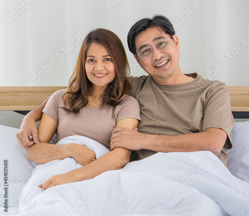 closeup of a happy middle age Asian husband and wife sitting on a bed together while a man put his arm around the shoulder of a woman smiling at a camera © Bangkok Click Studio