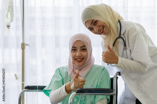 Young female muslim doctor talking and cheering up young sick muslim pateint sitting on wheelchair in hospital photo