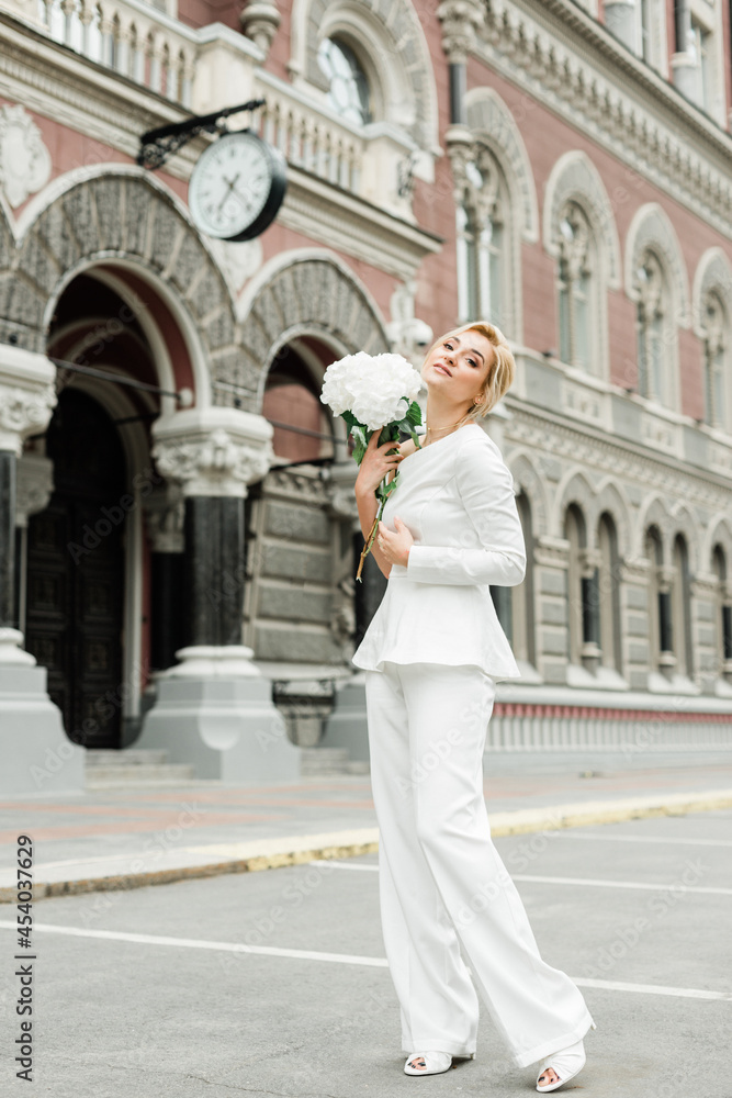 A young stylish lady in a white pantsuit with an elegant hairstyle with a bouquet of hydrangea flowers on the streets of the city. The woman is the bride. Soft selective focus