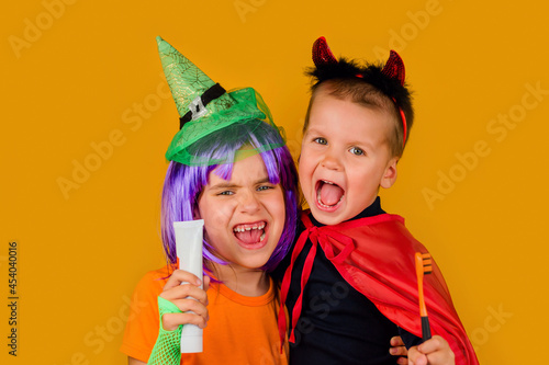 Two little toddler boy and girl in a carnival costume with toothpaste and an orange brush for Halloween is isolated on a yellow background. Medicine, dental hygiene, holidays concept.