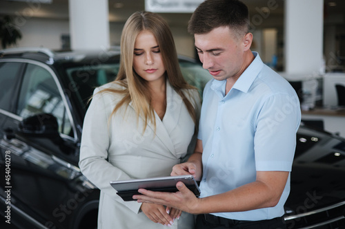 Salesperson talking with woman and help choosing new car. Man and woman in car showroom. Man using tablet to shop opportunity of different models