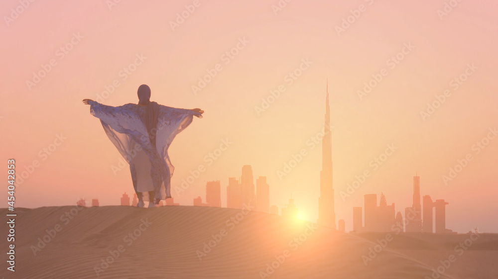 Φωτογραφία Arabic woman weared in traditional UAE dress - abayain rising her hands on the sunset at a desert with Dubai city silhouette on the background