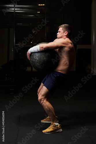 a handsome, muscular young man with a naked torso in a gym gradually lifts a huge stone ball from the floor. The ball has already been raised high enough. side view series multiple photos