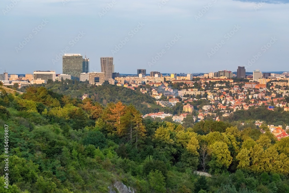 Prague, Czech Republic - August 21 2021: View of the city from distance with buildings and a skyscraper. Green trees in the foreground. Sunny summer evening, blue sky.