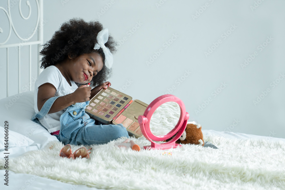 Little African girl playing makeup with mother's cosmetics. Adorable kid painting her lips with red lipstick and looking in the mirror with joy in bedroom at home Little African girl playing makeup with mother's cosmetics. Adorable kid painting her lips with red lipstick and looking in the mirror with joy in bedroom at home