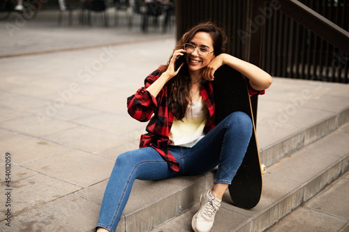 Portrait of young beautiful girl with skateboard. Happy smiling woman talking to the phone.