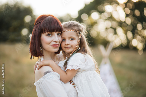 Close up portrait of happy beautiful mother and little daughter, posing at camera in the nature. Mom and child wearing white dress and feathers in hair, pretend to be native american