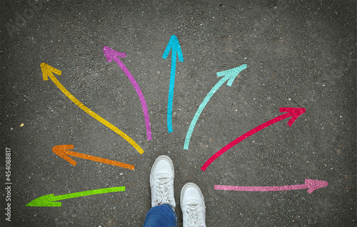 Legs in white sneakers in front of colorful arrows pointing in different directions on the gray asphalt. The concept of open opportunities and path choices photo