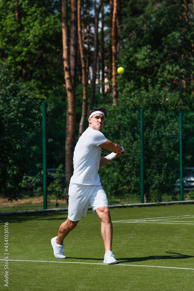 Focused sportsman in white sportswear playing tennis near ball on court