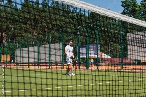 Young sportsman holding tennis racket near blurred net on court
