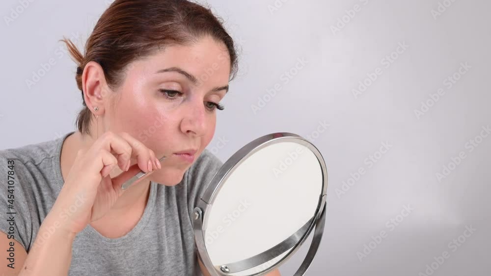 Caucasian woman looks in the mirror and removes the mustache herself with the help of tweezers. Hirsutism. Caucasian woman looks in the mirror and removes the mustache herself with the help of tweezers. Hirsutism.