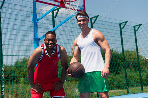 Cheerful african american sportsman looking at camera near friend with basketball ball