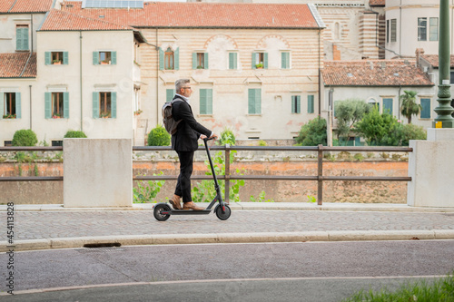 Side view full length of young businessman in a suit riding an electric scooter while commuting to work in city. Ecological transportation concept