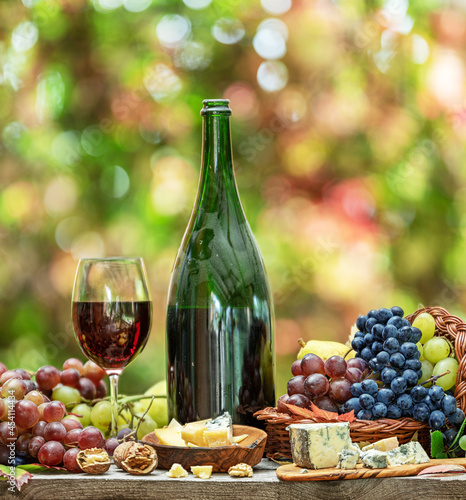 Grapes, bottle of wine and different cheeses on country wooden table and blurred colorful autumn background. Variety of products as the symbol of autumn abundance and prosperity.