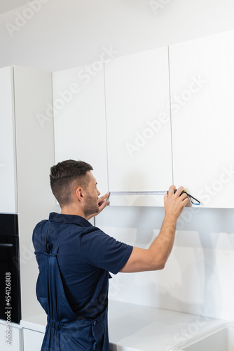 handyman in overalls measuring white furniture in kitchen