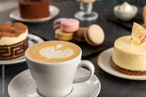 Café  con arte latte en un primer plano acompañado de distintos tipos de tortas con foco diferenciado.