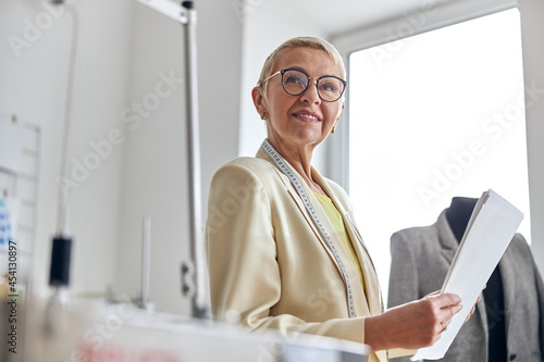 Thoughtful mature lady tailor holds papers in light sewing workshop