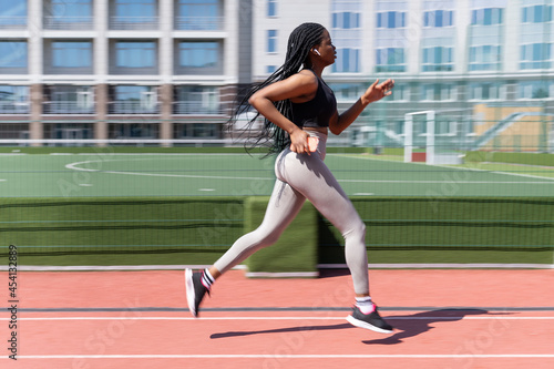 Young african woman running outdoors in stadium at summer day, motion blur. Fit sport girl jogging on athletic field. Runner afro female playing sports, hold smartphone and wear wireless headphones. 