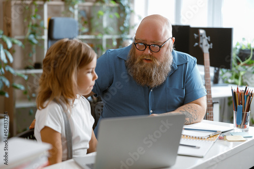 Bearded father and doubting little daughter at video lesson via laptop at home
