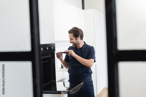 Fotografie smiling handyman repairing inbuilt electric oven with screwdriver on blurred for