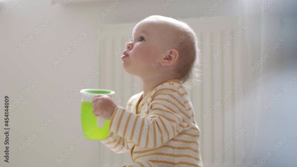 Portrait drinking baby at home. Child learns to drink on his own. Kid sitting on floor with plastic green cup in hands . Healthy food, lunch time. Portrait drinking baby at home. Child learns to drink on his own. Kid sitting on floor with plastic green cup in hands . Healthy food, lunch time.