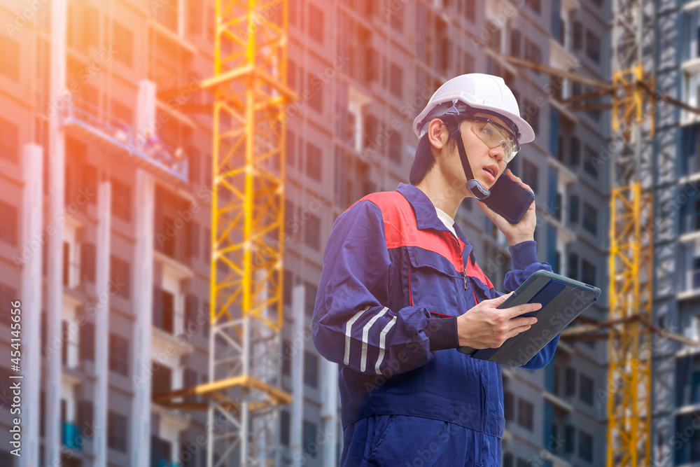 Low angle view of Asian engineer holding computer tablet and talking on smartphone while working in construction site area Low angle view of Asian engineer holding computer tablet and talking on smartphone while working in construction site area