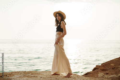 Young stylish woman standing on cliff at the sea at sunset