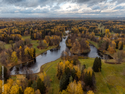Fototapeta Naklejka Na Ścianę i Meble -  Autumnal forest with golden crowns of trees. Colorful public park with lake. Foliage of trees change color from green to yellow and orange. Promenade. Cloudy sky. Change of season concept. Aerial view