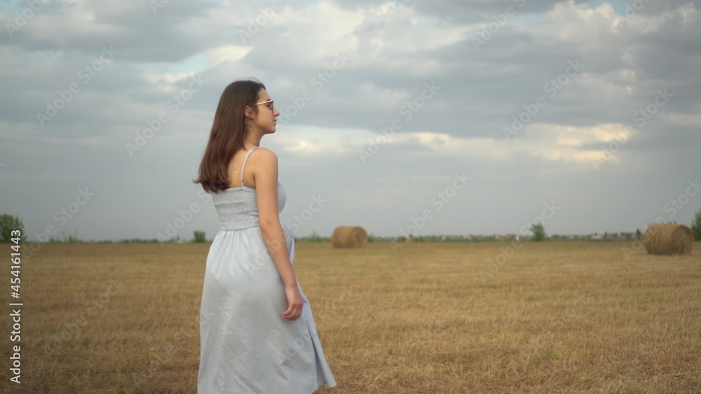 A young woman walks in a field with haystacks. Girl with glasses and a blue dress. Back view. A young woman walks in a field with haystacks. Girl with glasses and a blue dress. Back view.
