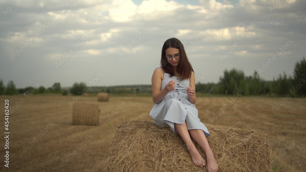 Young woman sits on a haystack in a field. A girl with glasses and a blue dress. Young woman sits on a haystack in a field. A girl with glasses and a blue dress.