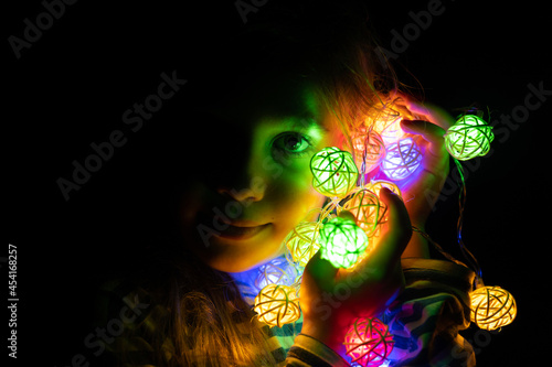 portrait of a little seven year old kid girl holding a garland in her hands near her face  glowing with colorful multicolored neon lights at night time at home. Christmas Eve holiday celebration