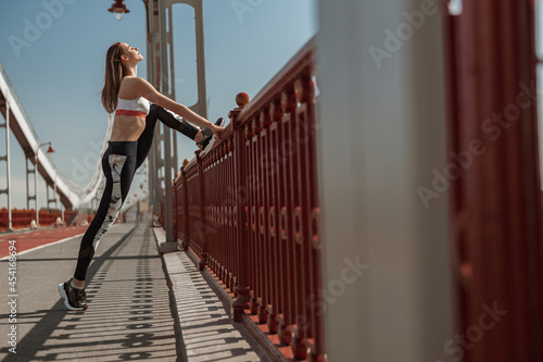 Tranquil slim woman stretches legs by handrail on emty footbridge